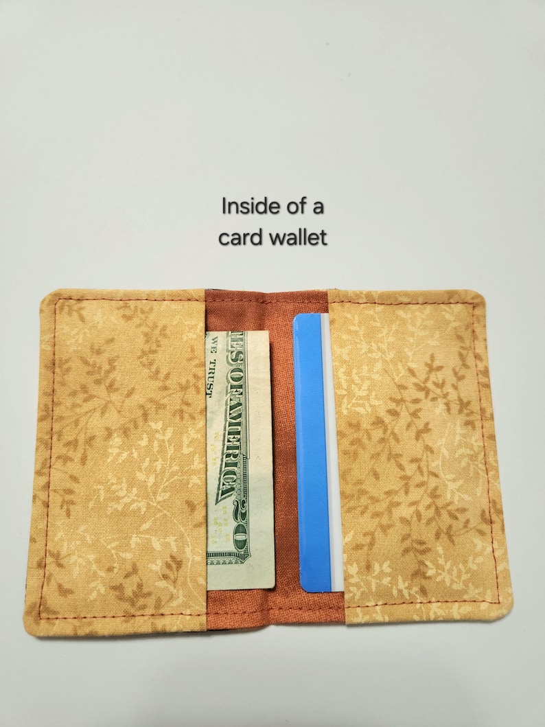 May include: A fabric card wallet with a tan and brown leaf pattern. The interior features two pockets containing a twenty-dollar bill and a blue card. The text 'Inside of a card wallet' is printed above the open wallet on a plain background.