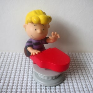 May include: A yellow-haired cartoon character playing a red toy piano. The character is wearing a purple and black striped shirt. The piano is on a gray base.