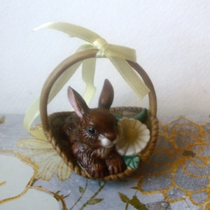 May include: A ceramic Easter bunny figurine in a woven basket. The brown bunny has long ears and is nestled with a white and yellow flower. A yellow ribbon is tied to the basket handle. The figurine is a decorative item.