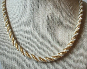 May include: A gold and white twisted rope necklace. The necklace is made of small beads and is on a fabric mannequin.