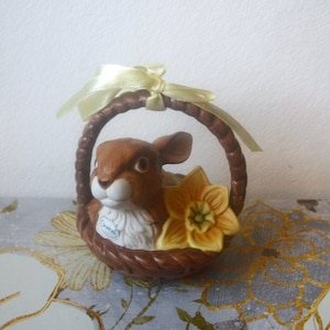 May include: A ceramic figurine of a brown bunny peeking out of a woven basket. The basket has a yellow ribbon tied to the handle and a yellow daffodil flower. The figurine is on a floral patterned surface.