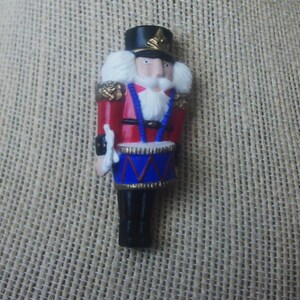 May include: A miniature, colorful nutcracker figurine. The figurine is dressed in a red jacket with gold accents, a black hat, and black boots. It has a white beard and holds a drum with blue and gold details. The figurine is approximately 3 inches tall.