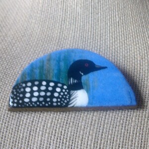 May include: A semi-circular brooch featuring a hand-painted common loon on a blue background. The bird has a black head with a red eye, a white neck patch, and a black body with white spots. The scene includes green reeds in the background.