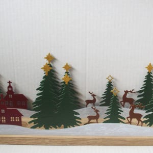 May include: A wooden Christmas decoration featuring a winter scene. The scene includes green fir trees topped with gold stars, brown reindeer, and a red church. The base is a light brown wood.