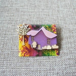 May include: A whimsical brooch featuring a purple house with a pink roof, set against a colorful background. A gold-colored tree is on the left. The brooch is square-shaped and has a glossy finish.