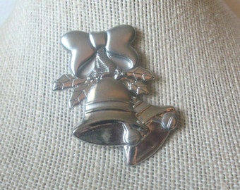 May include: A silver-toned metal brooch featuring two bells adorned with holly leaves and a bow at the top. The design has a polished, reflective surface with smooth curves and defined edges, showcasing a classic holiday-themed decorative accessory.