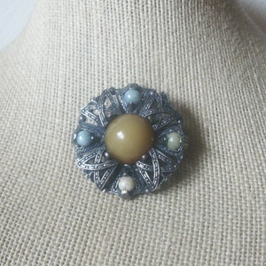 May include: A circular silver-toned brooch featuring an intricate openwork metal frame with a large, smooth, mustard-yellow central cabochon. Four smaller, round, multi-colored stone beads are set at the cardinal points within the metal design.
