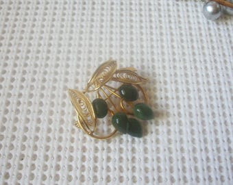 May include: A gold-tone brooch featuring delicate filigree metal leaves and four smooth, dark green, teardrop-shaped beads arranged to resemble a cluster of fruit. The intricate wirework creates an open, airy design with a polished finish.