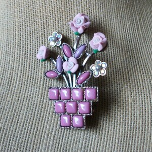 May include: A vintage brooch featuring a bouquet of pink roses and lavender leaves in a square, pink, mosaic-style vase. The brooch has silver-tone metal accents and small, white flower details. The brooch is a floral accessory.