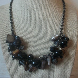 May include: A statement necklace featuring a dark metallic chain adorned with a cluster of polished black beads, irregular brown stone fragments, and shimmering grey shell-like pieces. The design creates a dense, textured arrangement of dark tones.