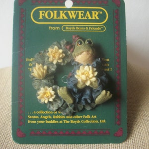 May include: A Folkwear figurine from Boyds Bears & Friends. The figurine is a green frog wearing a blue dress and a red hat, surrounded by a wreath of yellow flowers and green leaves. The packaging is green with red accents.