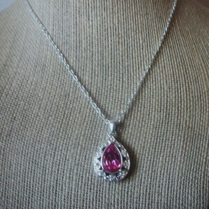 May include: A silver necklace with a teardrop-shaped pendant. The pendant features a large, vibrant pink gemstone surrounded by a halo of smaller, clear stones. The necklace is displayed against a neutral, textured background.