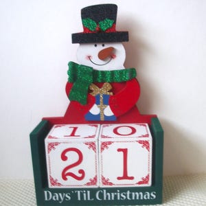 May include: A wooden countdown calendar featuring a snowman wearing a black glittery top hat with holly and a green glittery scarf. The snowman holds a blue gift with a gold bow. Two white cubes with red numbers sit in a green base labeled Days 'Til Christmas.