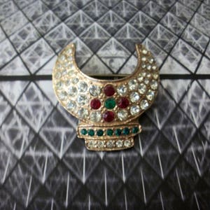 May include: A gold-toned brooch in a crescent moon shape, encrusted with clear and colored rhinestones. The brooch features a base with green and clear stones, and red and green stones in the center. The background is a black and white geometric pattern.