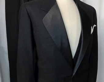 May include: A classic black tuxedo suit displayed on a mannequin. The suit features a satin lapel, a white pocket square, and a pair of matching black trousers. The suit is a formal wear ensemble, perfect for special occasions.