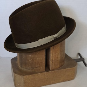 May include: A brown fedora hat with a light-colored band, displayed on a wooden hat block. The hat has a classic shape with a slightly indented crown and a brim. The hat block is made of light-colored wood.