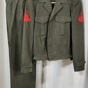May include: A green military uniform with a jacket and pants. The jacket has two large pockets on the front and a red patch on each sleeve. The pants have a red patch on the right leg.