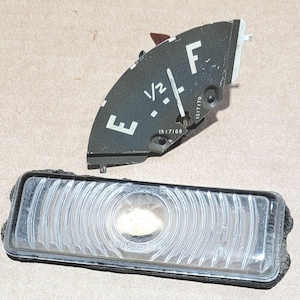 May include: A black and white fuel gauge with the letters "E" and "F" for empty and full. The gauge also has markings for 1/2 full. The gauge is missing a portion of the face. A clear plastic lens with a black frame is also shown.