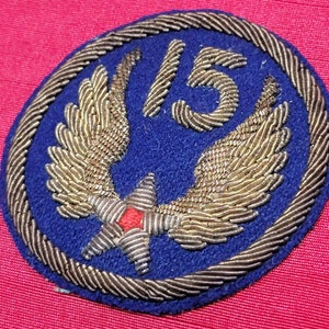 May include: A blue patch with gold-colored embroidered wings and the number 15. The patch has a gold-colored rope border and a red star at the bottom. The background is a solid red color.