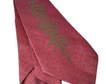 May include: A folded, burgundy-colored textile with a textured surface. A stylized, olive-green design is printed on the fabric. The textile is likely a scarf or a similar accessory.