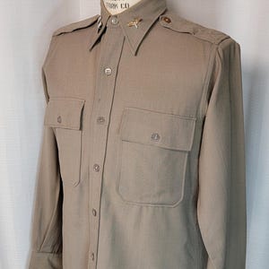 May include: A khaki long-sleeved shirt with a button-up collar, two chest pockets, and a gold pin on the collar. The shirt is on a mannequin.