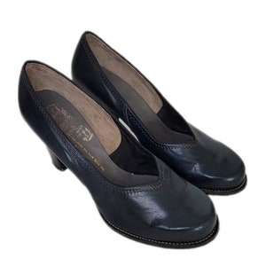 May include: Pair of dark blue leather heeled pumps with a pointed toe and a small v-shaped opening at the top. The shoes have a stitched seam around the edge of the sole.