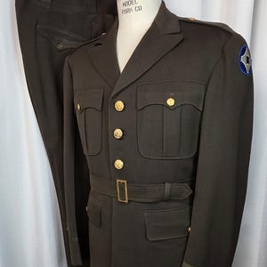 May include: A vintage olive drab military uniform, including a jacket with gold buttons and a belt, and matching trousers. The jacket features a patch on the shoulder. The uniform is displayed on a mannequin.