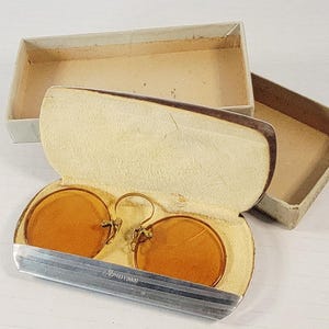 May include: Antique round amber-tinted eyeglasses with a gold-tone frame, resting in an open silver case with a cream-colored interior. The case is next to a matching cardboard box. The case has the text "PATENTED" on it.