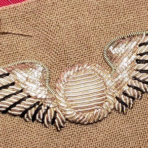 May include: A gold embroidered pilot's wings patch with a central circle, sewn onto a brown fabric background.