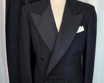 May include: A black double-breasted tuxedo jacket with satin lapels and matching trousers. A white pocket square is in the breast pocket. The suit is displayed on a mannequin. The trousers are hanging on a hanger.