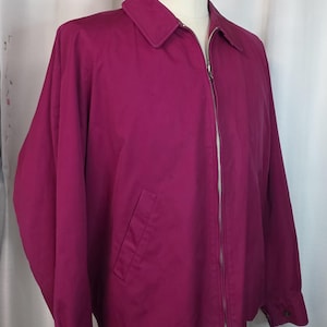 May include: A magenta-colored jacket with a full-length zipper, a collar, and a single pocket on the left side. The jacket is displayed on a mannequin. The sleeves have buttoned cuffs. The jacket is made of a smooth fabric.