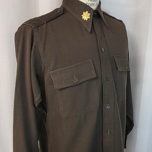 May include: A dark brown long-sleeved shirt with a button-up collar and two flap pockets. The shirt has a gold star pin on the collar. The text "SUPERIOR MODEL FORK CO" is visible on the collar.