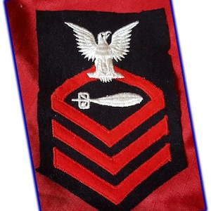 May include: A black and red embroidered patch featuring a white eagle above a red and black emblem with a bomb shape. Below the emblem are three red chevrons on a black background. The patch is on a red fabric.