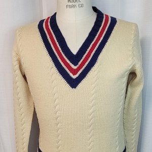 May include: A cream-colored cable knit sweater with a V-neck and navy and red trim. The sweater has a label that reads "Superior Model Fork Co."