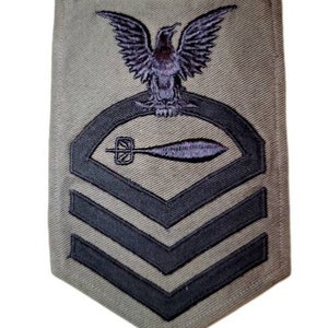 May include: A green fabric patch with a black eagle and a black torpedo. The patch has three black stripes on the bottom.