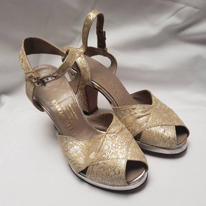 May include: A pair of gold and silver open-toe heeled sandals with a floral pattern. The sandals have a buckle closure and the brand name "FRANCO FERRER" is printed on the insole.