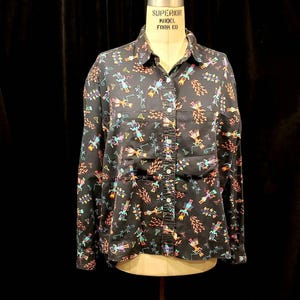 May include: A black long-sleeved button-down shirt with a colorful geometric pattern. The shirt has a collar and a button-up front. The pattern features stylized figures and symbols in shades of blue, green, orange, and red.