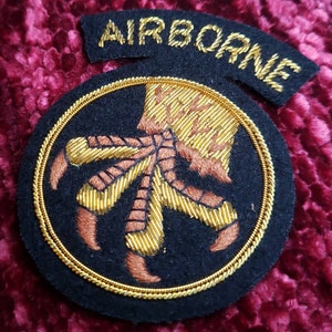 Peut inclure: Écusson brodé noir avec une bordure dorée et le mot "AIRBORNE" en arc de cercle en haut. L'écusson présente un motif de griffe doré et marron détaillé au centre.