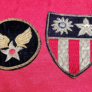 Peut inclure: Deux écussons brodés sur fond rouge. L'un est rond avec des ailes dorées et une étoile rouge sur du velours noir. L'autre est en forme de bouclier avec un dessus bleu, des étoiles argentées et des rayures rouges et argentées.