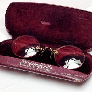 May include: A pair of round, pink-tinted glasses in a burgundy leather case. The case has the inscription "Columbian Optical Co. Seattle 1315 4th Ave."
