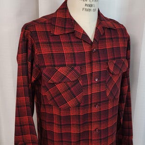 May include: A red and black plaid long-sleeve shirt with a button-up front and two chest pockets. The shirt has a collar and is made of a soft, comfortable fabric. The label on the shirt reads "Superior Model Form Co."