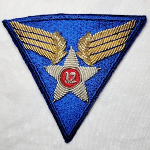 Peut inclure: Un écusson triangulaire bleu avec des ailes brodées en or et une étoile argentée avec un cercle rouge contenant le chiffre "12" au centre.