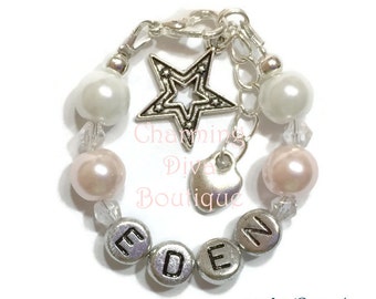 May include: A silver charm bracelet with white and light pink pearls, silver beads, and a silver star charm. The bracelet also has a silver heart charm and silver letter beads that spell out the name "EDEN". The bracelet is labeled "#9 Lt Peach".