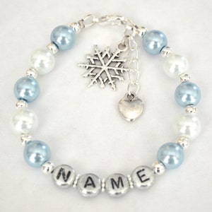 May include: A blue and white beaded bracelet with a silver snowflake charm and a heart charm. The bracelet has the name "NAME" spelled out in silver beads.