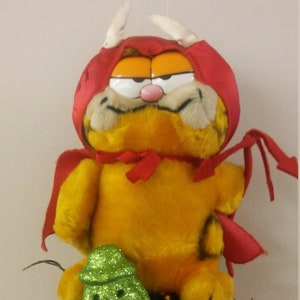 May include: A red and orange plush Garfield cat wearing a red devil costume sits atop a red Christmas ornament. The ornament is decorated with black and gold tinsel, a green glitter pumpkin, and a white ghost.