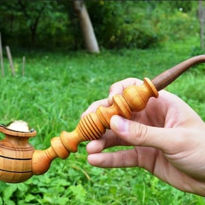 May include: A wooden smoking pipe with a round bowl and a long, curved stem. The pipe is made of light brown wood and has a brass lid on the bowl.