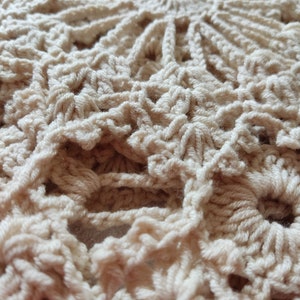 May include: Close-up of a cream-colored, crocheted doily. The handmade piece features a circular design with intricate patterns and openwork details. The texture of the yarn is visible, creating a soft and inviting aesthetic. The doily is likely used for home decor.