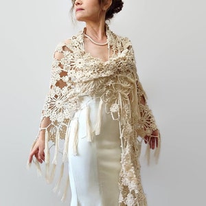 May include: Cream-colored, open-weave crochet shawl with floral and geometric patterns. The shawl features long fringe and is draped over a white dress. The design is intricate and delicate, suitable for special occasions.