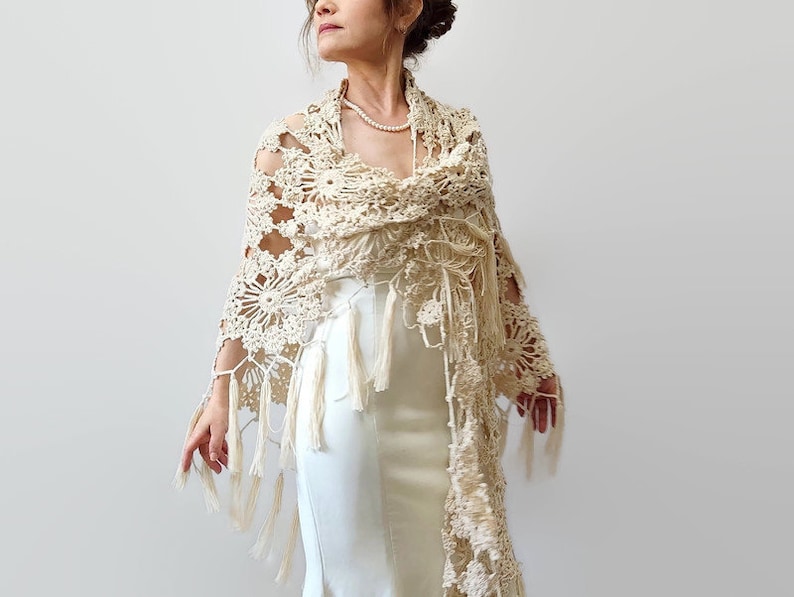 May include: Cream-colored, open-weave crochet shawl with floral and geometric patterns. The shawl features long fringe and is draped over a white dress. The design is intricate and delicate, suitable for special occasions.