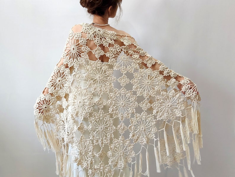 Cream-colored, open-weave crochet shawl with a floral pattern and fringe trim. Scialle all'uncinetto color crema a trama aperta, con motivo floreale e finiture a frange. Châle au crochet de couleur crème, à maille ajourée, orné d'un motif floral et de franges. Cremefarbenes Häkeltuch in grober Netzoptik mit Blumenmuster und Fransenbesatz. Chal de ganchillo de color crema y tejido calado, con motivo floral y remate de flecos.